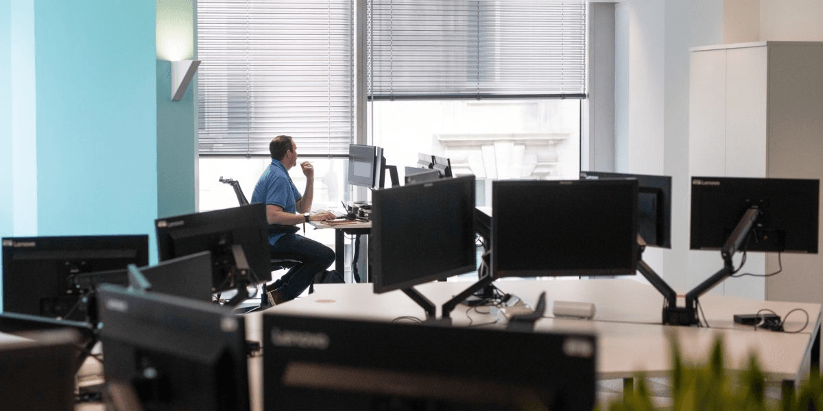 Mostly empty open office with rows of unoccupied workstations and one person working alone at a desk by the window.